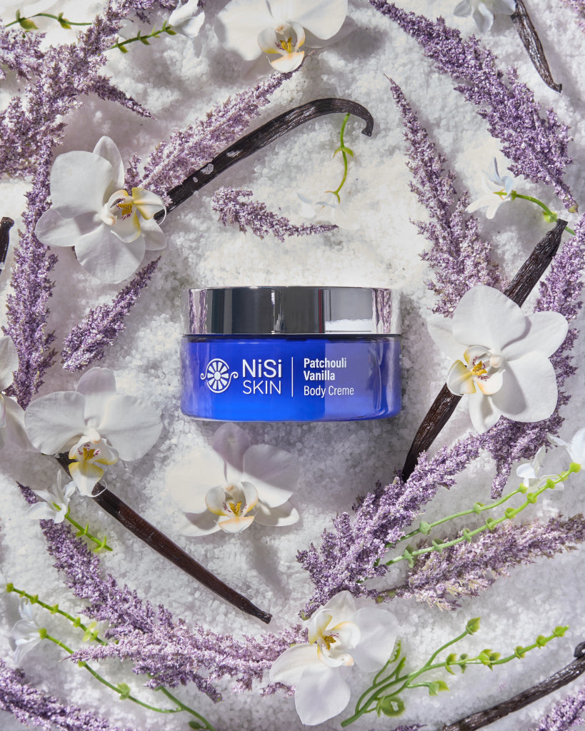 Nisi Skin Patchouli vanilla body creme in a blue jar with a metal lid displayed in an assortment of raw materials including vanilla purple and white flowers