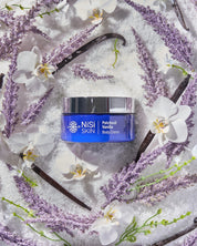 Nisi Skin Patchouli vanilla body creme in a blue jar with a metal lid displayed in an assortment of raw materials including vanilla purple and white flowers