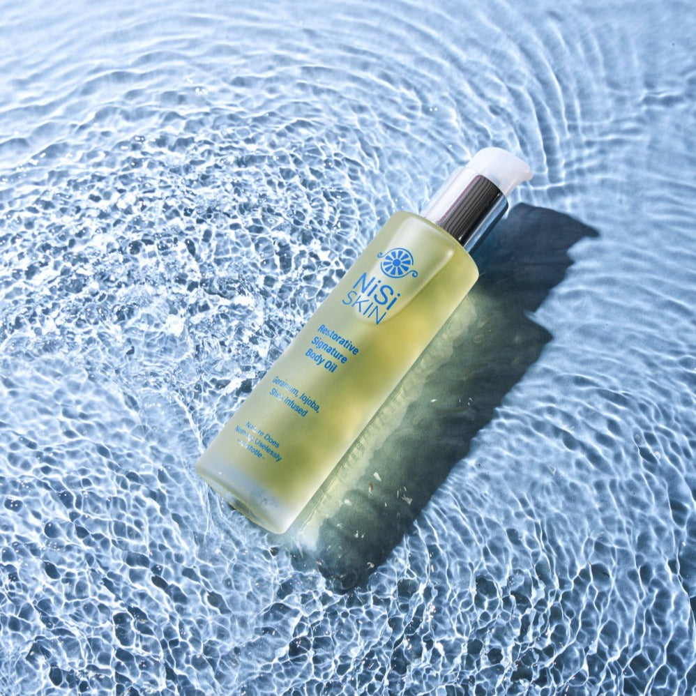 Nisi Skin Restorative Signature Body oil golden liquid geranium golden oil in glass bottle in pool of blue rippled water reflecting light